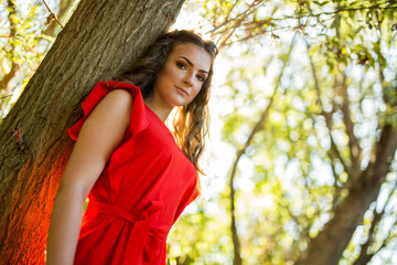 Poster - Shallow focus shot of a Caucasian woman wearing makeup and a red dress leaning on a tree