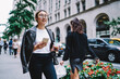 Attractive female blogger with mobile technology walking at street during free time in urban city, beautiful hipster girl in spectacles holding coffee to go and cellular device exploring town