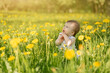 A little girl sits in a field of yellow dandelions in the sun and looks up to the side. The cute kid stuck his fingers in his mouth. Baby care. Licking dirty hands is dangerous for your health.