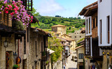 Fototapeta Uliczki - Perspective view of houses lined up in narrow street of old town and church in the background. Santillana del Mar.