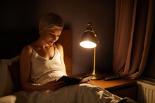 Beautiful female in pajamas lying on bed reading book, alone at night. Caucasian short haired lady in bedroom, charming cute woman in room lighted by lamp