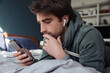 © Anna Tabakova/Stocksy - Man looks at his phone wearing headphones in bed