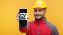 Delivery Man Wearing Yellow Helmet And Uniform Showing Mobile Phone To The Camera With QR Code On The Screen. Yellow Background Studio Shot. Digital Pay Concept. 