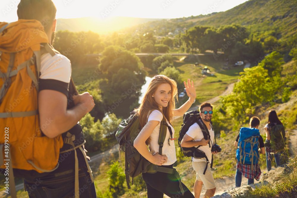 Group of happy backpackers trekking on sunny day. Young tourists traveling and enjoying active summer vacation. Smiling woman looking at camera and waving hand walking down hiking trail with friends - obrazy, fototapety, plakaty 