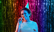 birthday, celebration and lgbt concept - happy smiling young african american woman in party cap with blower in ultraviolet neon lights over foil fringe curtain in rainbow colors background