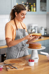 Wall Mural - culinary, baking and cooking food concept - happy smiling young woman making layer cake on kitchen at home