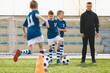 © matimix - Coaching Kids Soccer. Young Man as Soccer Coach Trainer on Football Children Camp. Soccer Boys in Blue Uniforms Running Fast and Kicking Balls on Training Pitch