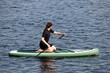 Girl in black wetsuit with paddle kneeling down on a board in a water. Standup paddleboarding, summer leisure