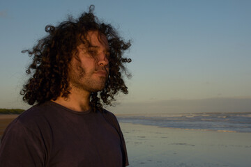 portrait of a curly young Caucasian man on the beach. He looks towards the camera with his hair blowing in the wind.  curly hair for men isolated with the sea in the background. very lush male hair.