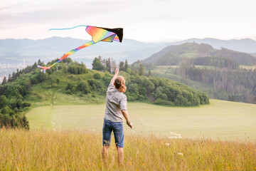 Wall Mural - Teenager boy on the green hills meadow grass launching colorful rainbow kite toy with a long tail. Happy childhood moments or outdoor time spending concept image.