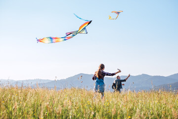 Wall Mural - Little daughter with the father running fast while they flying up colorful kites on the high grass meadow in the mountain fields. Warm family moments or outdoor time spending concept image.