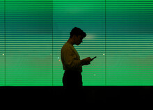 Young Non-binary Person Standing In Front Of Green Lighted Screen With Smartphone