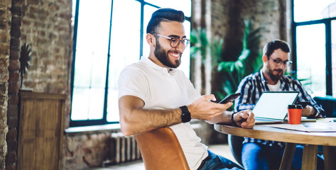 Happy Turkish IT professional enjoying smartphone chatting during collaborative meeting with remote colleague for programming on laptop, cheerful hipster guy in eyewear reading funny text publication