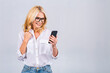 Happy mature senior woman holding smartphone using mobile online apps, smiling old middle aged lady texting sms message chatting on phone looking at cellphone isolated on white grey background