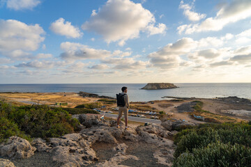 Man with backpack is hiking along a rocky coast at sunset near the Mediterranean sea in Cyprus. Male tourist admires the view Yeronisos, or Holy Island, lies off the coast of Agios Georgios Pegeias