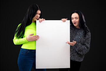 two attractive funny young women with a white blank poster, isolated on dark background