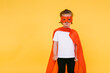 Little blonde girl dressed as a superheroine superhero with a cape and red mask, angry, with the cape to the side, on yellow background