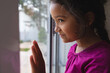 © Wavebreak Media - Happy hispanic girl standing at window with hand on glass, looking out and smiling