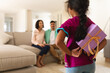 © Wavebreak Media - Hispanic parents sitting on couch with daughter standing in foreground hiding present behind back
