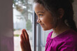 © Wavebreak Media - Happy hispanic girl standing at window with hand on glass, looking out and smiling