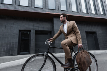 young businessman in formal wear holding leather brown bag and riding bicycle outside.