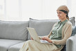 Candid portrait of mature woman using headscarf and using laptop while relaxing on couch in cozy home setting, copy space