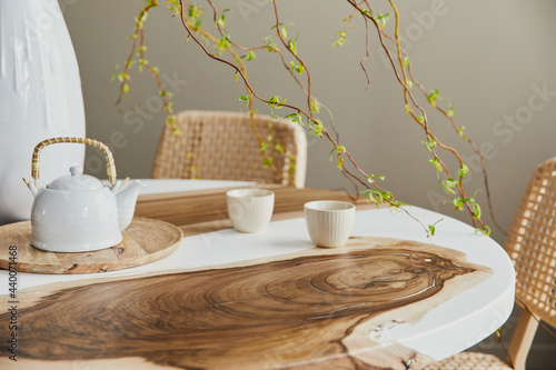 Interior design of stylish dining room interior with family wooden and epoxy table, rattan chairs, flowers in vase and teapot with cups.  Details. Template.