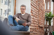© Studio 27/Westend61 - Mature man in eyeglasses using digital tablet while sitting on window sill at home