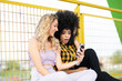 © Jose Carlos Ichiro/Westend61 - Surprised Afro woman looking at smart phone while sitting with female friend by fence
