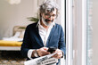 © COROIMAGE/Westend61 - Smiling senior man with newspaper using smart phone near window at home