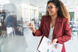 © SERGIO NIEVAS/Westend61 - Smiling businesswoman holding financial report while writing on glass in office
