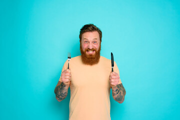Wall Mural - Happy man with tattoos is ready to eat with cutlery in hand