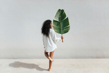 Curly Haired Woman Holding Big Banana Leaf While Standing On One Leg In Front Of White Wall During Sunny Day