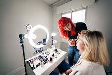Female Make-up Artist With Dyed Hair Applying Eyeshadow On Model While Filming Through Mobile Phone At Studio