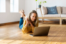 Blond Woman With Credit Card Lying Down On Floor While Using Laptop At Home