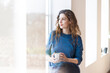© steve brookland/Westend61 - Young woman holding coffee mug while sitting at window in living room