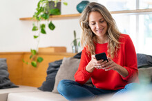 Smiling Blond Woman Text Messaging On Mobile Phone While Sitting In Living Room At Home