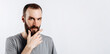 © Daria Lukoiko - Serious puzzled handsome male model with beard holding hand on chin as if thinking about something, squinting at camera with suspicious look and standing over gray background. Man decides what to buy