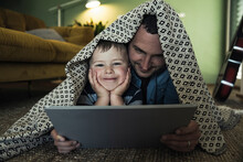 Smiling Father And Son With Digital Tablet Lying Under Blanket In Living Room