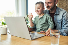 Happy Boy Waving Hand While Sitting With Father Doing Video Call Through Laptop At Home