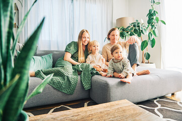 Happy young family of four relaxing on a couch, couple playing with baby girl and toddler boy