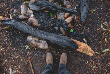 Guy Legs And Feet With Leather Shoes Over The Ground With Old Extinguished Campfire With Burned Wood In The Chilean Patagonia In A Rainy Day