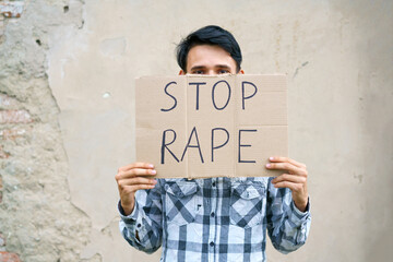 Young man holding cardboard with the inscription stop rape. Guy of Caucasian appearance against violence holds a poster with a protest at the demonstration against the background of the wall