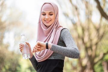 Wall Mural - Pleasant woman in hijab looking at smart tracker for checking jogging distance. Young female athlete in sport clothing standing at green park and holding bottle of water.