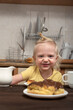 Cheerful blonde child in kitchen is having breakfast and looking at the cakes. Little girl drinking tea with sweets