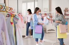 Happy Pregnant Women With Shopping Bags Choosing Baby Clothes In Store