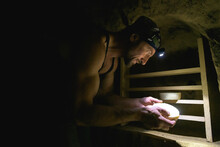 Side View Of Adult Farmer With Headlamp Looking At Handmade Cheese In Cellar 