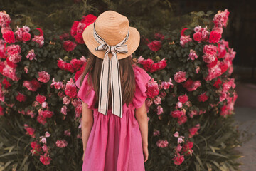 Poster - Cute child girl 5-6 year old wear straw hat with bow and summer dress posing with garden roses outdoors. Summer season. Childhood. Stylish toddler over nature background. Back view.