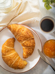 On a beautiful white tablecloth we see a table set for breakfast. On a white earthenware plate, two croissants, orange jam, a cup of coffee. There is a table knife on the plate.