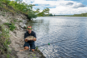 Wall Mural - Feeder fishing. Fisherman with big bream fish in hands and tackle at wild river shore